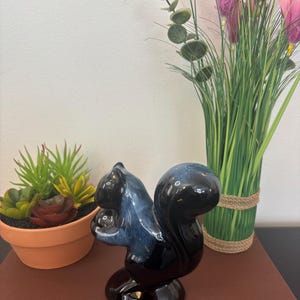 以下が含まれることがあります： 光沢のある濃い青と黒の仕上げが施されたセラミック製のリスの置物。 リスはナッツを持っており、丸い台座の上にいます。 置物は、鉢植えの多肉植物と装飾的な花と一緒に展示されています。