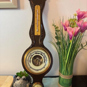May include: A dark brown wooden barometer with a thermometer and a circular gauge. The thermometer displays Fahrenheit readings. The gauge has a gold-colored frame and a white face with black text. The barometer is a decorative weather instrument.