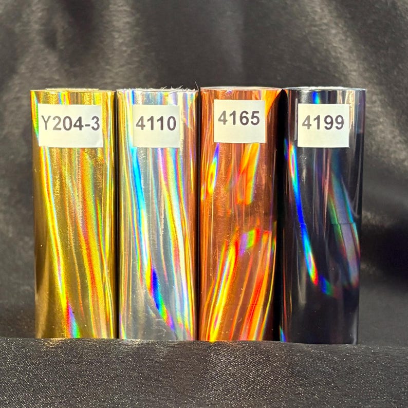 May include: Four rolls of holographic vinyl in gold, silver, copper, and black. Each roll has a white label with a product code. The vinyl reflects a rainbow of colors when light hits it, creating a shimmering effect.