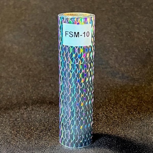 May include: A cylindrical roll of holographic film featuring a repeating silver scale pattern that reflects rainbow colors. A white rectangular label is affixed to the top portion of the roll, clearly displaying the black printed text FSM-10.