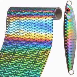 May include: A roll of holographic film featuring a repeating iridescent fish scale pattern in shades of green, blue, and gold, positioned next to a metal fishing lure with an identical scale texture and a prominent red and black eye detail.
