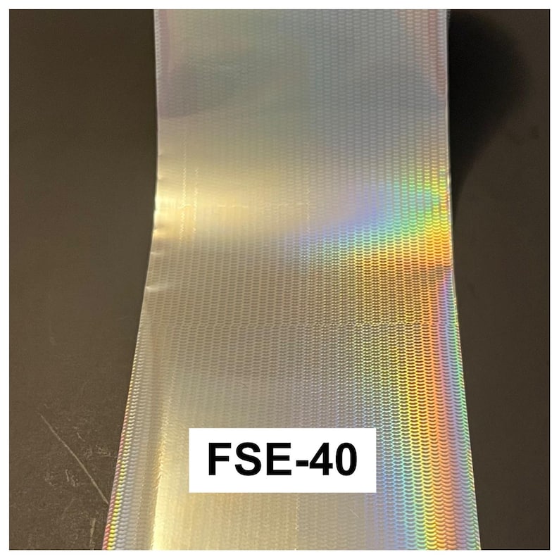 May include: A roll of silver holographic foil featuring a repeating pattern of small, horizontal, wave-like indentations. The surface reflects a spectrum of rainbow colors when light hits it. A white rectangular label displays the text FSE-40.