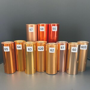 Może przedstawiać: Kolekcja metalicznych rolek folii w r&oacute;żnych odcieniach złota, miedzi i brązu. Każda rolka jest oznaczona białą naklejką z numerem. Rolki są ułożone w rzędzie, prezentując swoje r&oacute;żne kolory i wykończenia.