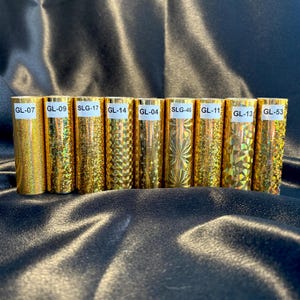 May include: A collection of gold-colored, cylindrical objects with varying textures and patterns. Each cylinder has a white label with a unique alphanumeric code, such as "GL-07" or "SLG-17". The objects are arranged in a row against a dark, reflective background.