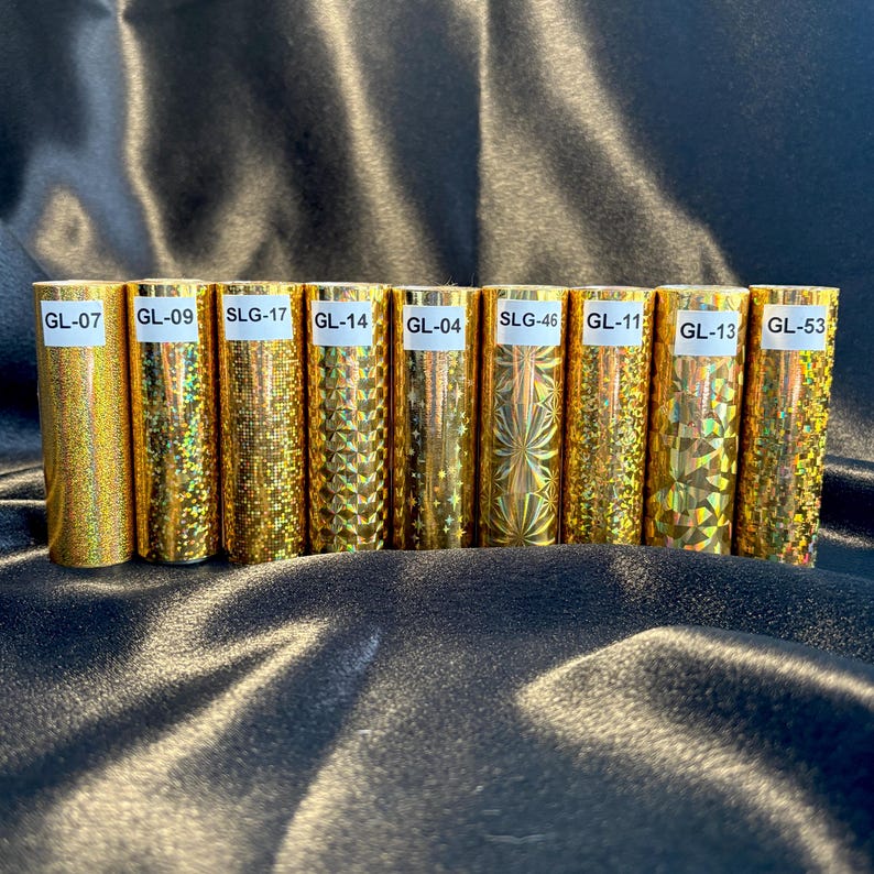 May include: A collection of gold-colored, cylindrical objects with varying textures and patterns. Each cylinder has a white label with a unique alphanumeric code, such as "GL-07" or "SLG-17". The objects are arranged in a row against a dark, reflective background.