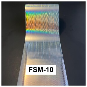 May include: A roll of silver holographic transfer foil featuring a repeating fish scale pattern. The surface reflects a spectrum of rainbow colors when light hits it. A white rectangular label with the black text FSM-10 is visible on the foil.