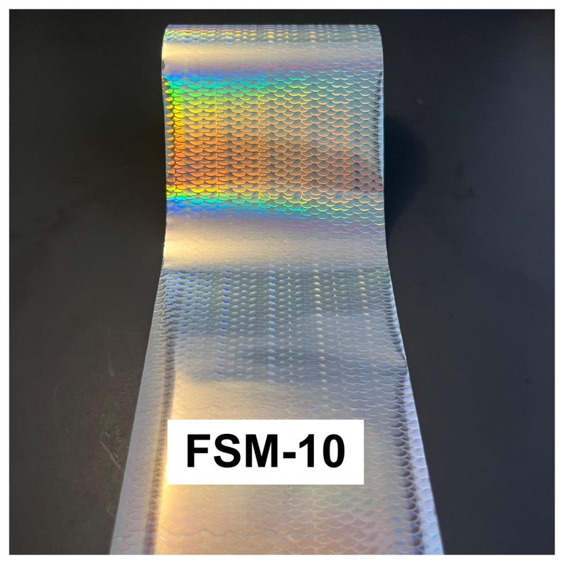 May include: A roll of silver holographic transfer foil featuring a repeating fish scale pattern. The surface reflects a spectrum of rainbow colors when light hits it. A white rectangular label with the black text FSM-10 is visible on the foil.