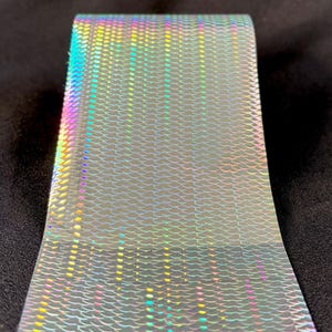 May include: A roll of holographic transfer foil featuring a repeating fish scale or mermaid tail pattern. The surface reflects a spectrum of iridescent colors including blue, green, yellow, and pink, creating a shimmering effect across the material.