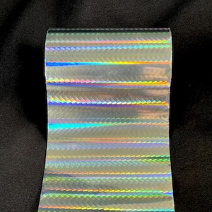 May include: A roll of holographic transfer film featuring a repeating chevron pattern that reflects a spectrum of rainbow colors. The surface of the material is smooth and metallic, displaying shifting light effects across the horizontal bands.