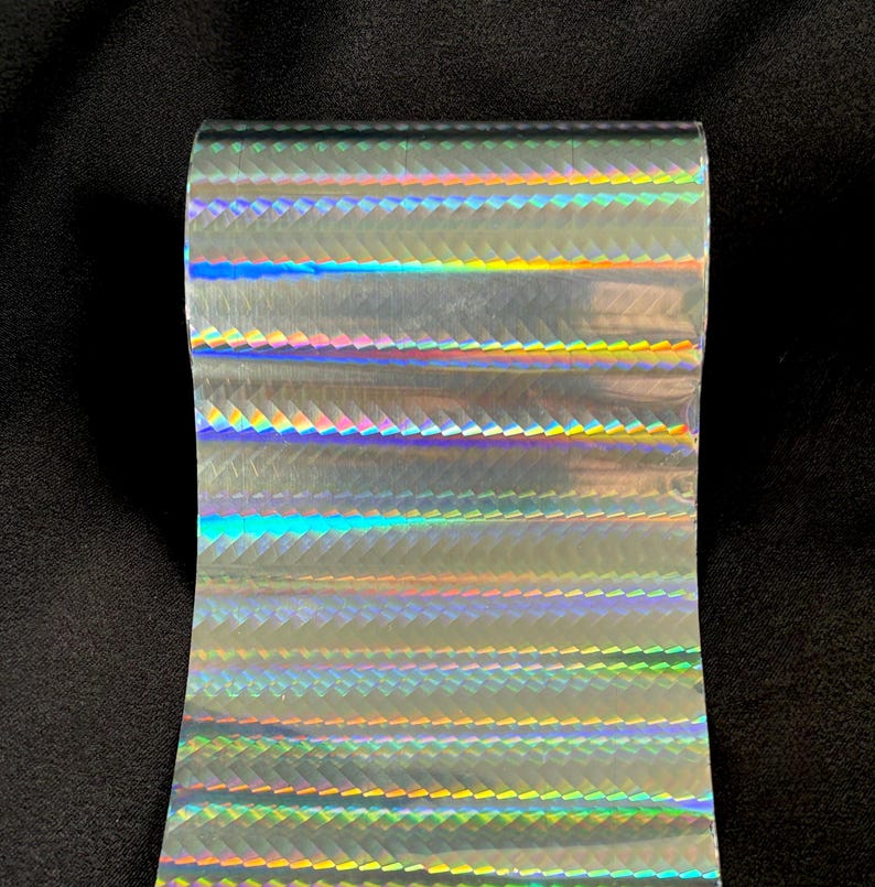 May include: A roll of holographic transfer film featuring a repeating chevron pattern that reflects a spectrum of rainbow colors. The surface of the material is smooth and metallic, displaying shifting light effects across the horizontal bands.