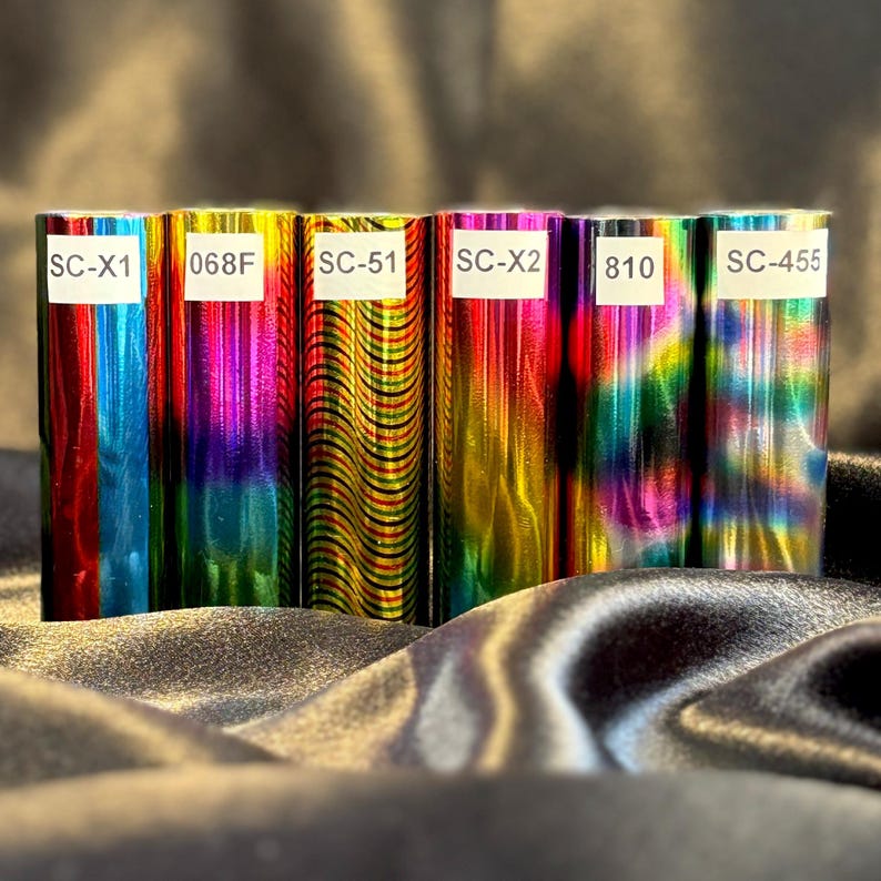 May include: A collection of six rolls of colorful, metallic vinyl. Each roll displays a vibrant array of colors, including blues, greens, pinks, and yellows. White labels with black text, such as "SC-X1" and "068F", are affixed to the rolls. The rolls are arranged on a dark, textured surface.
