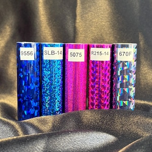 May include: Five rolls of decorative foil in shades of blue, pink, and iridescent. Each roll has a unique pattern and a white label with a number and letter combination. The foil is displayed on a dark, textured surface.