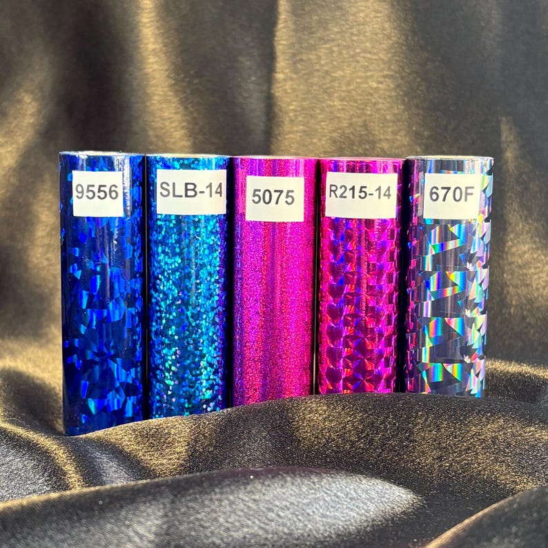 May include: Five rolls of decorative foil in shades of blue, pink, and iridescent. Each roll has a unique pattern and a white label with a number and letter combination. The foil is displayed on a dark, textured surface.