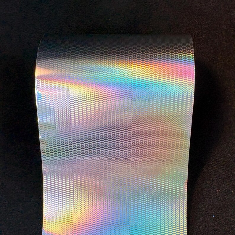 May include: A roll of holographic vinyl material featuring a repeating pattern of small, interlocking semicircular scales. The surface reflects a spectrum of iridescent colors, shifting from warm yellows and oranges to cool blues and purples.