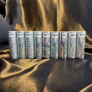 May include: A collection of ten cylindrical rolls with holographic designs. Each roll is labeled with a unique code, such as "S2-8" and "5710". The rolls are silver and reflect light, creating a shimmering effect. The background is a dark, textured fabric.