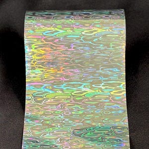 May include: A roll of transfer foil featuring a holographic, iridescent water ripple pattern. The surface displays shifting colors including green, blue, pink, and yellow, creating a reflective, multi-dimensional effect across the entire length of the material.