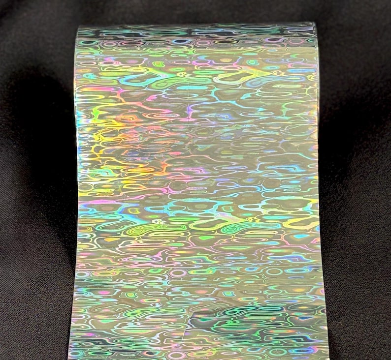 May include: A roll of transfer foil featuring a holographic, iridescent water ripple pattern. The surface displays shifting colors including green, blue, pink, and yellow, creating a reflective, multi-dimensional effect across the entire length of the material.