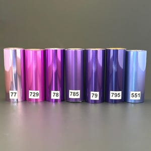 Może przedstawiać: Siedem rolek folii metalicznej w odcieniach fioletu i niebieskiego. Każda rolka ma białą etykietę z numerem: 77, 729, 78, 785, 79, 795 i 551. Folia ma błyszczącą, odblaskową powierzchnię.