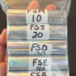 May include: A clear plastic resealable bag containing five rolls of silver holographic craft vinyl. Each roll features a unique texture and is labeled with white stickers marked FSM 10, FSJ 20, FSD 30, FSE 40, and FSB 50 in black ink.