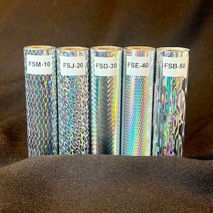 May include: Five cylindrical rolls of holographic foil featuring distinct reflective patterns. Each roll has a white label with black text: FSM-10, FSJ-20, FSD-30, FSE-40, and FSB-50. The patterns include scales, concentric circles, diagonal lines, and waves.