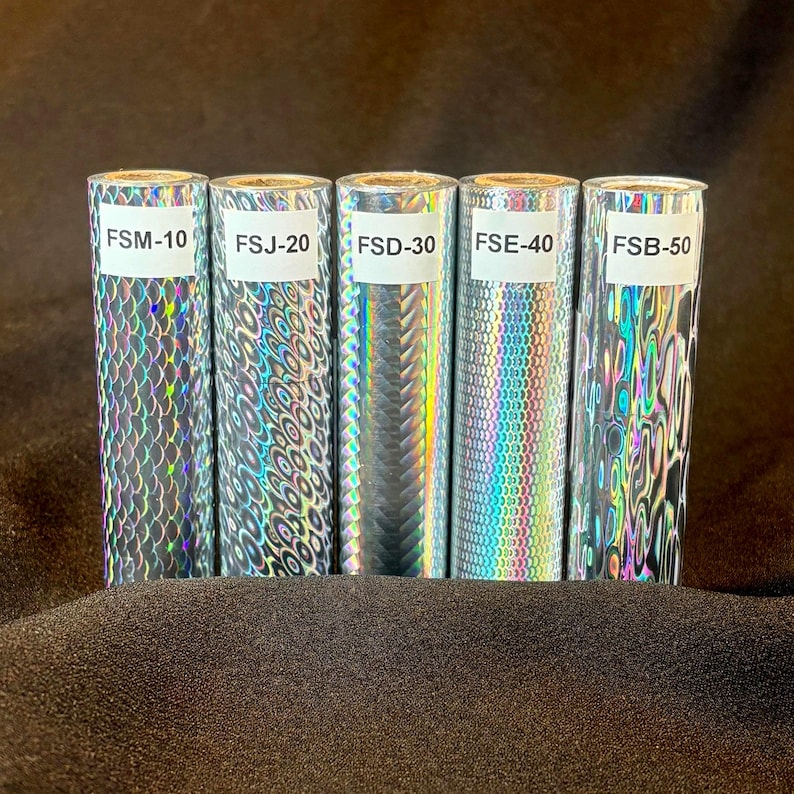 May include: Five cylindrical rolls of holographic foil featuring distinct reflective patterns. Each roll has a white label with black text: FSM-10, FSJ-20, FSD-30, FSE-40, and FSB-50. The patterns include scales, concentric circles, diagonal lines, and waves.