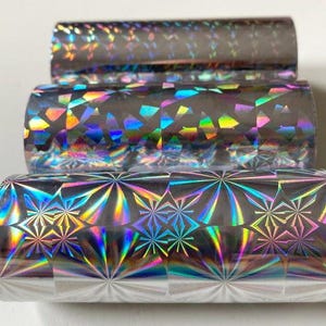 May include: Three rolls of holographic foil in a stack. The foil has a shiny, reflective surface with a rainbow of colors. The top roll has a subtle pattern, the middle roll has a geometric pattern, and the bottom roll has a starburst design.