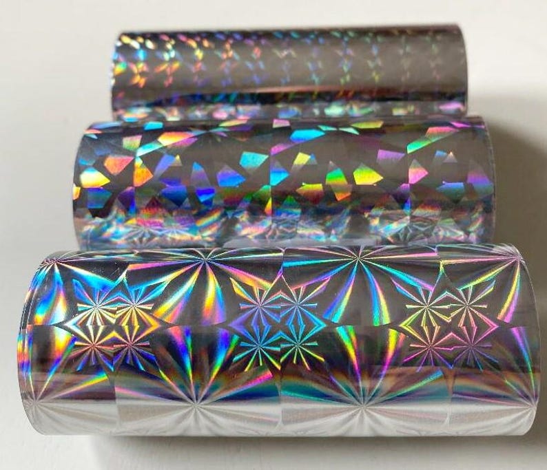 May include: Three rolls of holographic foil in a stack. The foil has a shiny, reflective surface with a rainbow of colors. The top roll has a subtle pattern, the middle roll has a geometric pattern, and the bottom roll has a starburst design.