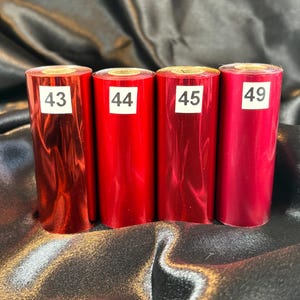 Może przedstawiać: Cztery rolki czerwonej folii z białymi etykietami z numerami 43, 44, 45 i 49. Folia ma błyszczący, metaliczny wygląd. Rolki są ułożone pionowo na czarnej, teksturowanej powierzchni.