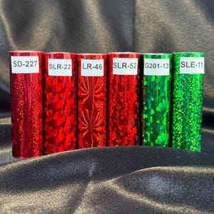 May include: Six rolls of decorative foil in red and green. Each roll has a unique pattern and is labeled with a white sticker. The labels read: SD-227, SLR-22, LR-46, SLR-52, G201-13, and SLE-11. The foil has a shiny, reflective surface.