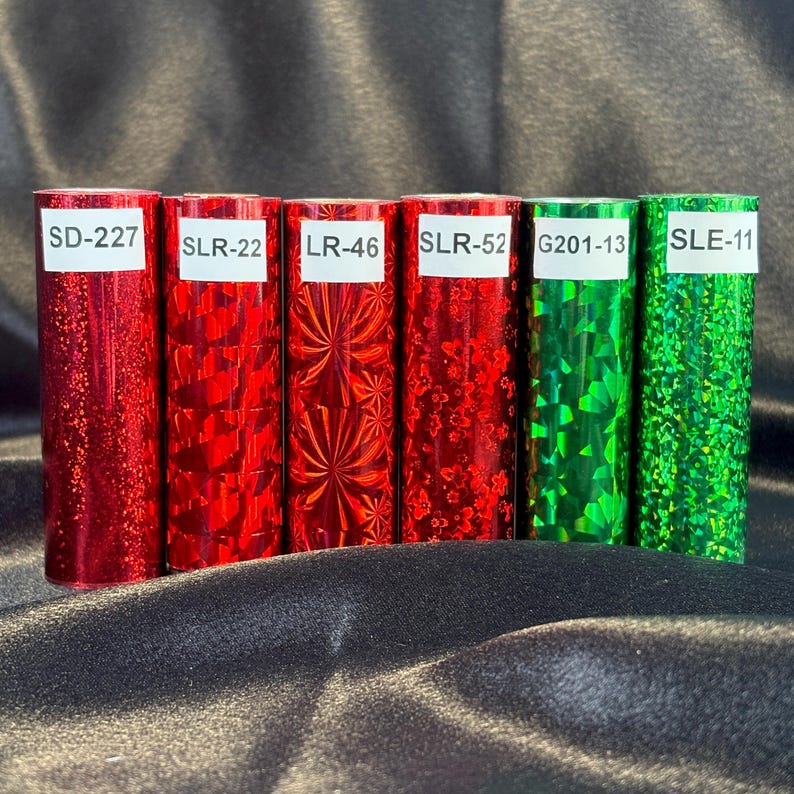 May include: Six rolls of decorative foil in red and green. Each roll has a unique pattern and is labeled with a white sticker. The labels read: SD-227, SLR-22, LR-46, SLR-52, G201-13, and SLE-11. The foil has a shiny, reflective surface.