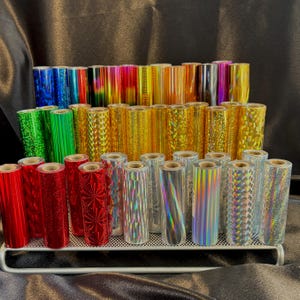 May include: A collection of colorful foil rolls in various patterns and colors, including red, green, gold, and silver. The rolls are arranged on a tiered metal display, showcasing a variety of textures and designs. The foil appears to be for crafting or decorative purposes.