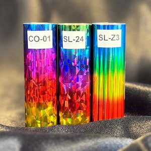 May include: Three rolls of iridescent foil with a geometric pattern. Each roll is a different color, including blue, purple, red, and yellow. The rolls have white labels with the codes "CO-01", "SL-24", and "SL-Z3".