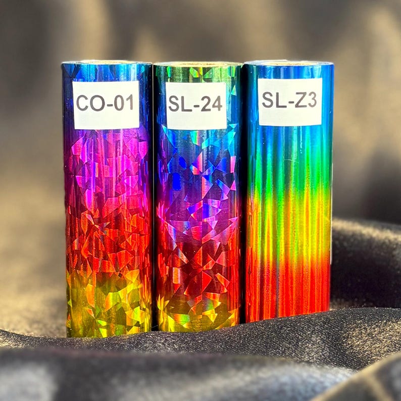 May include: Three rolls of iridescent foil with a geometric pattern. Each roll is a different color, including blue, purple, red, and yellow. The rolls have white labels with the codes "CO-01", "SL-24", and "SL-Z3".