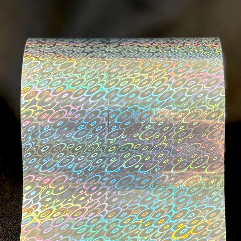 May include: A roll of adhesive tape featuring a repeating holographic pattern of overlapping circles. The surface reflects a spectrum of iridescent colors including blue, green, yellow, and pink, creating a shimmering effect across the entire material.