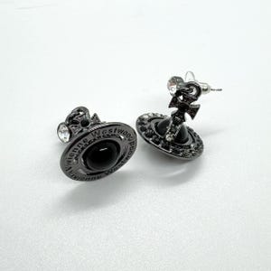 May include: A pair of dark gray stud earrings. Each earring features a black orb with a cross design and is encircled by small, clear crystals. The brand name "Vivienne Westwood" is inscribed around the orb.