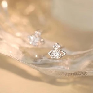 May include: A pair of small, silver-toned stud earrings. Each earring features a central, round, clear gemstone, surrounded by a decorative silver setting. The earrings are set against a soft, blurred background.