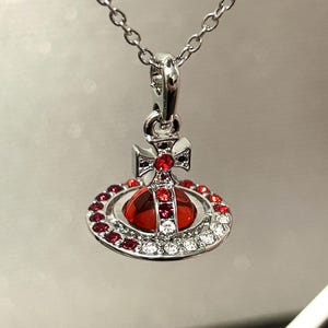 May include: Silver-toned pendant necklace featuring a red orb design with a cross detail. The orb is encircled by red and clear crystals, suspended from a delicate chain. A stylish accessory.