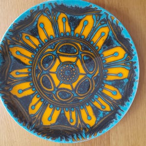 Op de afbeelding: Een ronde keramische schaal met een levendig bloemmotief. De schaal heeft een turkooizen rand, een zwarte achtergrond en een centraal geel bloemmotief met blauwe accenten. Waarschijnlijk een vintage stuk.