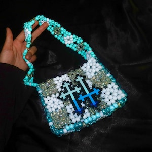 May include: A handmade handbag constructed from plastic pony beads in shades of turquoise, white, and iridescent clear. The bag features a repeating geometric pattern and two prominent cross-shaped motifs in dark blue and light blue gradient tones.