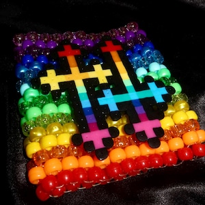 Op de afbeelding: Een brede armband gemaakt van rijen kleurrijke plastic kralen in een regenboogverloop. In het midden van de armband bevinden zich twee zwarte kruisvormige bedels, elk met een verticaal regenboogkleurig streeppatroon.