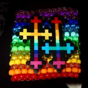 May include: A multi-row cuff bracelet made of vibrant, translucent plastic pony beads arranged in a rainbow color gradient. Two large, black-bordered cross shapes made of fused plastic beads are attached to the center, featuring a rainbow pattern.