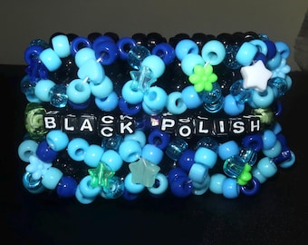 May include: A multi-layered cuff bracelet made of various shades of blue, black, and translucent plastic pony beads. The bracelet features star and flower-shaped beads and includes black square letter beads spelling out the words BLACK POLISH.
