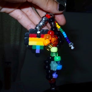 May include: A keychain featuring a black heart-shaped charm with horizontal rainbow stripes, accented by a collection of multi-colored plastic pony beads in red, orange, yellow, green, blue, purple, and black, strung on a metal ring.