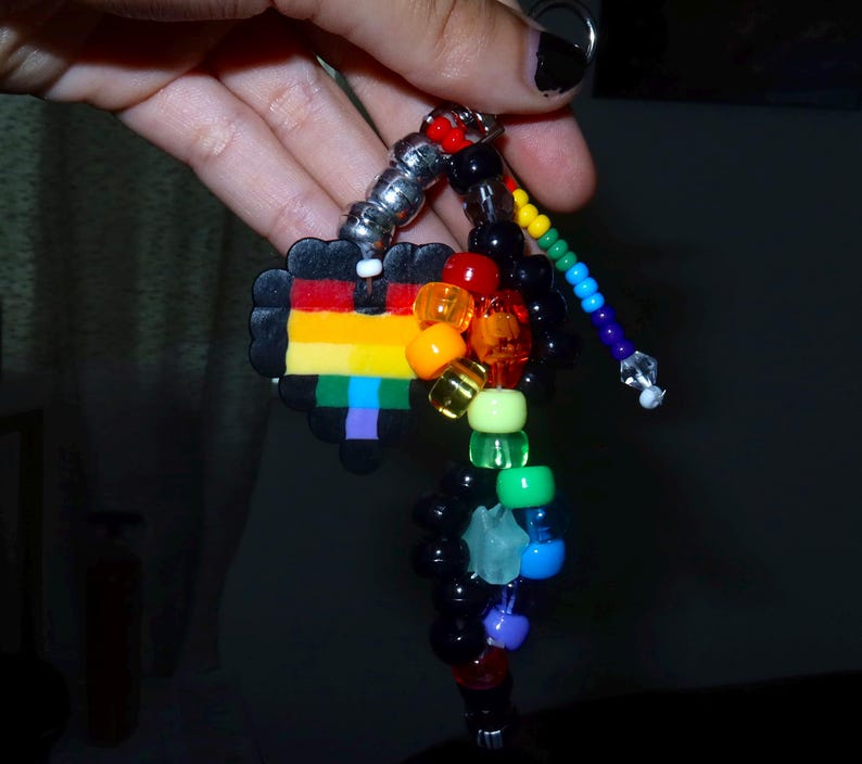 May include: A keychain featuring a black heart-shaped charm with horizontal rainbow stripes, accented by a collection of multi-colored plastic pony beads in red, orange, yellow, green, blue, purple, and black, strung on a metal ring.