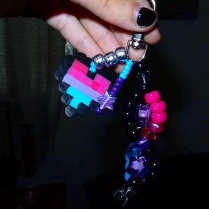 May include: A keychain featuring a pixelated heart charm made of black, pink, and blue beads. The charm is attached to a silver metal clasp with a strand of colorful plastic beads, including metallic silver, bright blue, purple, and hot pink spheres, along with a small purple star-shaped bead.