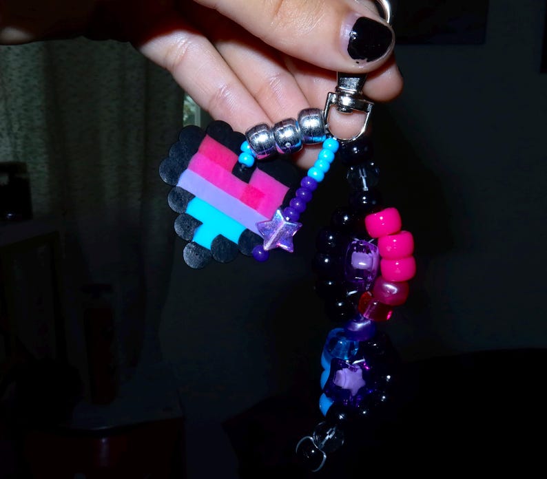May include: A keychain featuring a pixelated heart charm made of black, pink, and blue beads. The charm is attached to a silver metal clasp with a strand of colorful plastic beads, including metallic silver, bright blue, purple, and hot pink spheres, along with a small purple star-shaped bead.