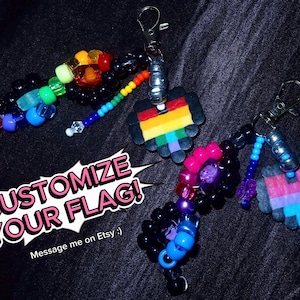 May include: Two keychains featuring black plastic beads, colourful pony beads, and pixelated heart charms attached to silver-tone lobster clasps. The text CUSTOMIZE YOUR FLAG! is displayed in white and pink lettering over a white speech bubble graphic.