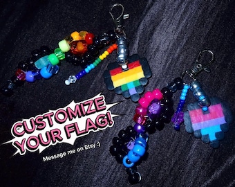 May include: Two keychains featuring black plastic beads, colorful pony beads, and pixelated heart charms attached to silver-tone lobster clasps. The text CUSTOMIZE YOUR FLAG! is displayed in white and pink lettering over a white speech bubble graphic.