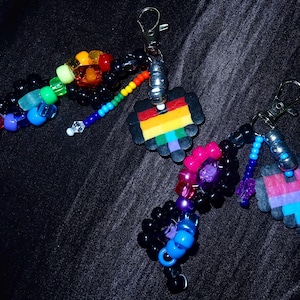 May include: Two keychains featuring metal lobster clasps, silver-toned beads, and pixelated heart charms. One keychain has a rainbow-striped heart with black, blue, and green beads. The other has a pink, purple, and blue heart with black and pink beads.