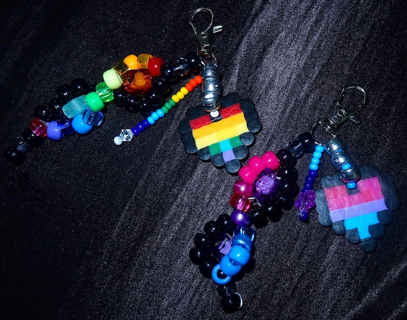 May include: Two keychains featuring metal lobster clasps, silver-toned beads, and pixelated heart charms. One keychain has a rainbow-striped heart with black, blue, and green beads. The other has a pink, purple, and blue heart with black and pink beads.